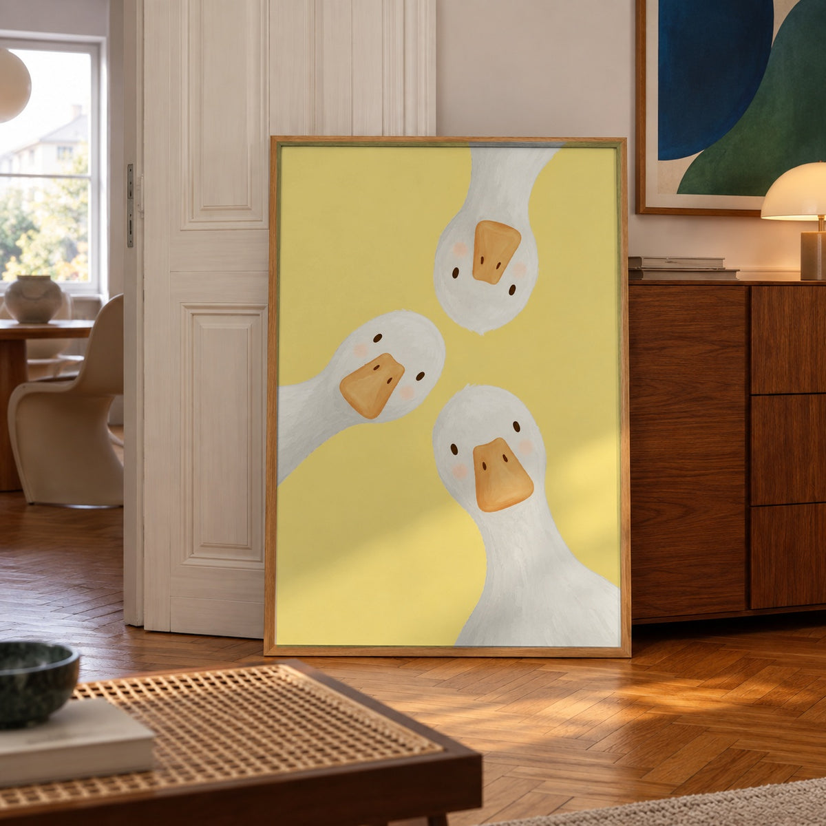 Three blushing-cheeked geese lean in close on a cheerful yellow background — poster displayed in a styled room