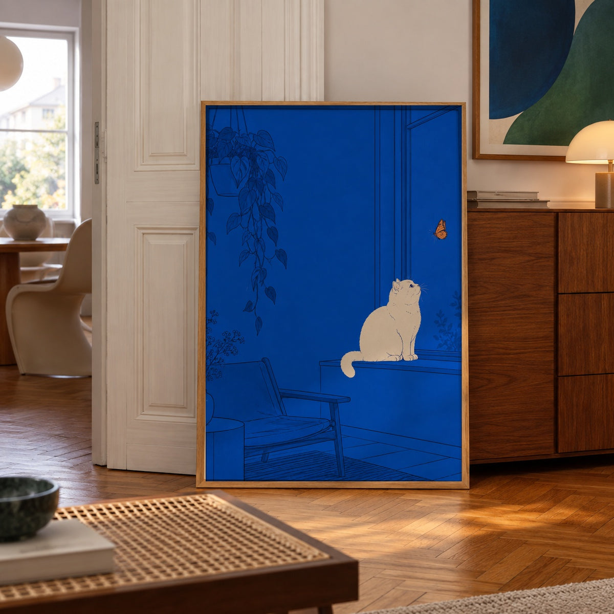A cream-colored cat perches on a windowsill, captivated by a monarch butterfly fluttering just beyond the glass — poster displayed in a styled room