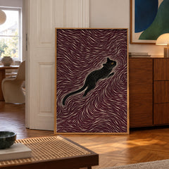 A sleek black cat stretches gracefully through a hypnotic swirl of maroon and cream lines — poster displayed in a styled room